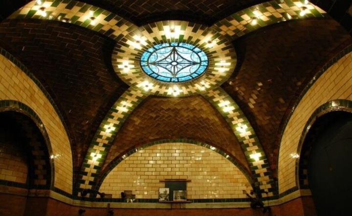 Historic New York subway station interior featuring a domed ceiling, arched walls covered in green and brown mosaic tiles, and a blue stained-glass skylight.