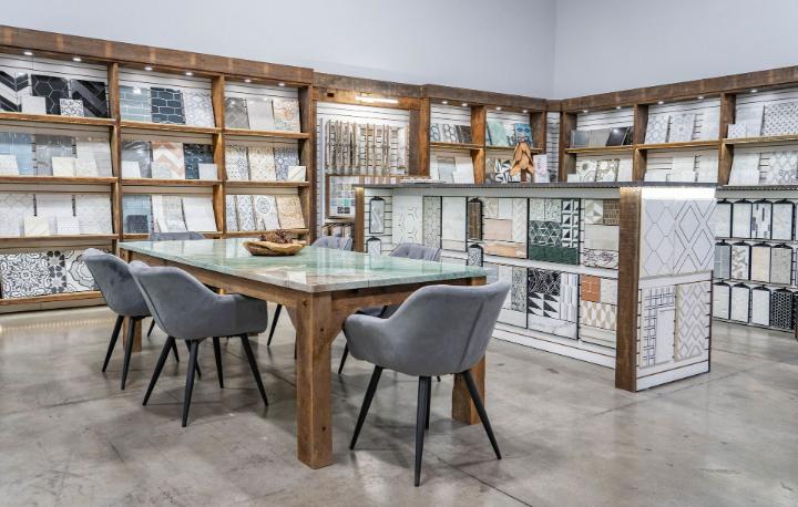 Tile showroom with shelves displaying diverse mosaic and ceramic tile styles, and a central table with gray chairs.